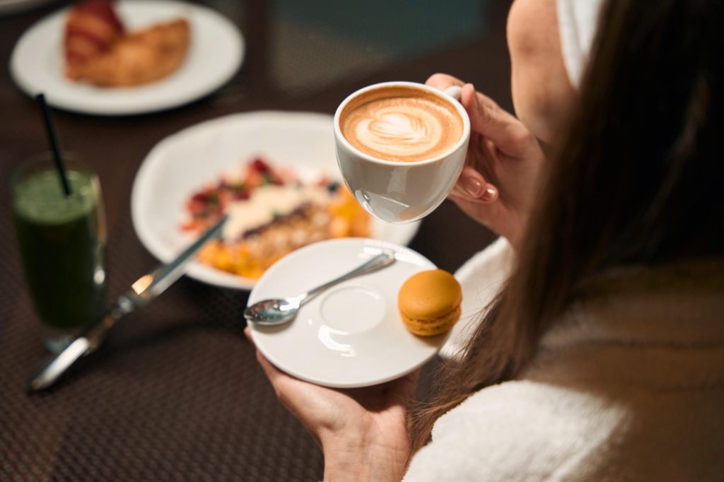 An adult woman holding a cup of coffee with a plate of macarons and a smoothie in a cozy breakfast setting.