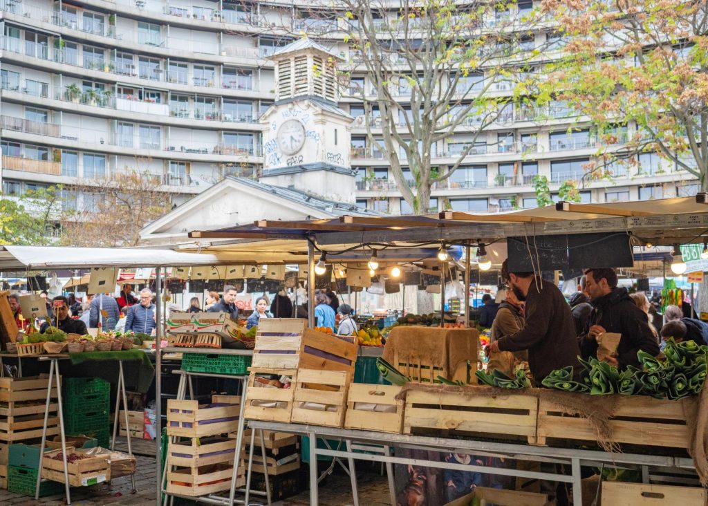 A bustling market scene at Aligre Market in Paris, featuring stalls with fresh produce, shoppers browsing, and a historic clock tower in the background.