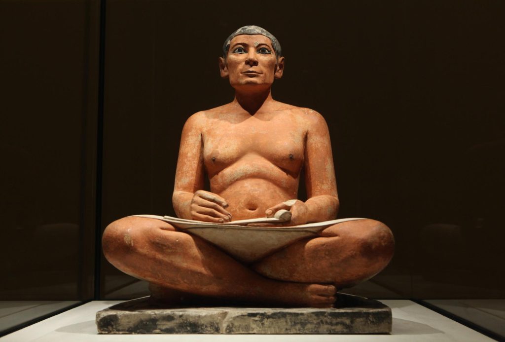 Ancient Egyptian sculpture of a sitting scribe, depicting a male figure in a seated position, holding a writing tablet and stylus.