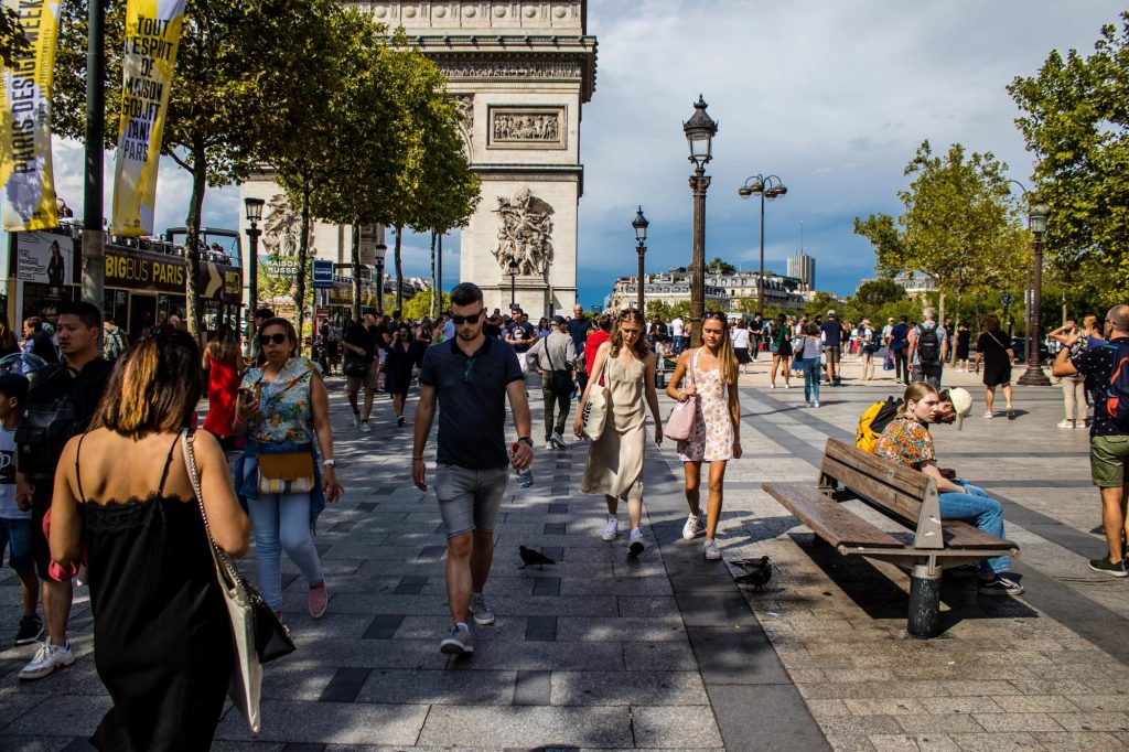 A busy street scene near the Arc de Triomphe in Paris, featuring pedestrians walking, people sitting on a bench, trees lining the walkway, and various shops and landmarks in the background.