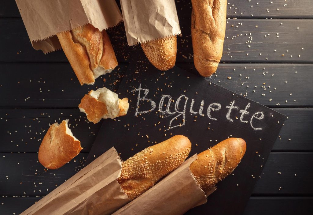 A variety of baguette bread, some in paper wrappers, with sesame seeds scattered around and the word 'Baguette' written in chalk on a black chalkboard.