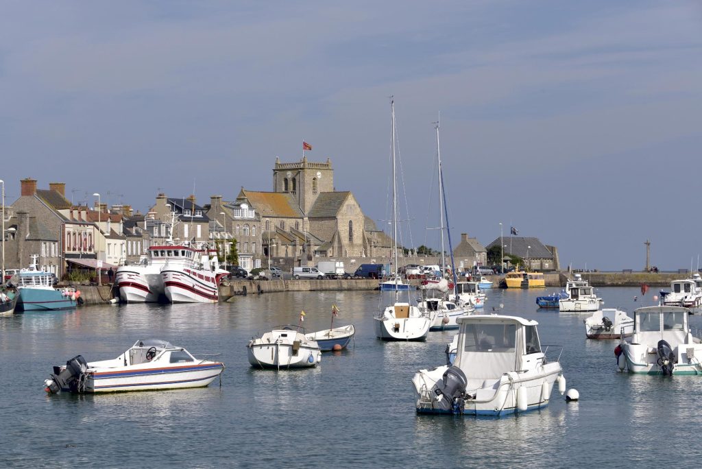 A picturesque view of Barfleur harbor with various boats docked in the calm water, surrounded by historic buildings and a church in the background.