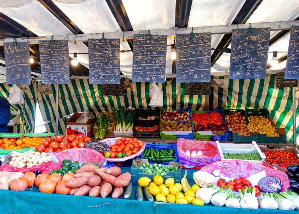 A colorful display of fresh fruits and vegetables at a market stall in Bastille, featuring tomatoes, sweet potatoes, lemons, and various greens.