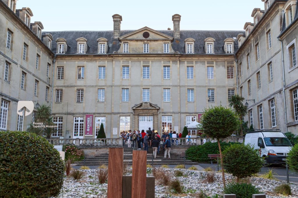 Entrance of the Bayeux Tapestry Museum with visitors gathering outside and a scenic courtyard.