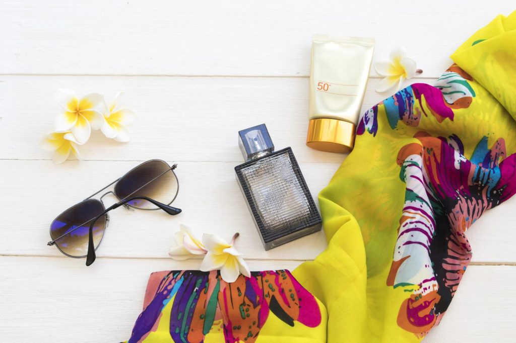 An arrangement of summer accessories including sunglasses, a floral scarf, perfume bottle, and sunscreen on a white wooden surface, adorned with tropical flowers.