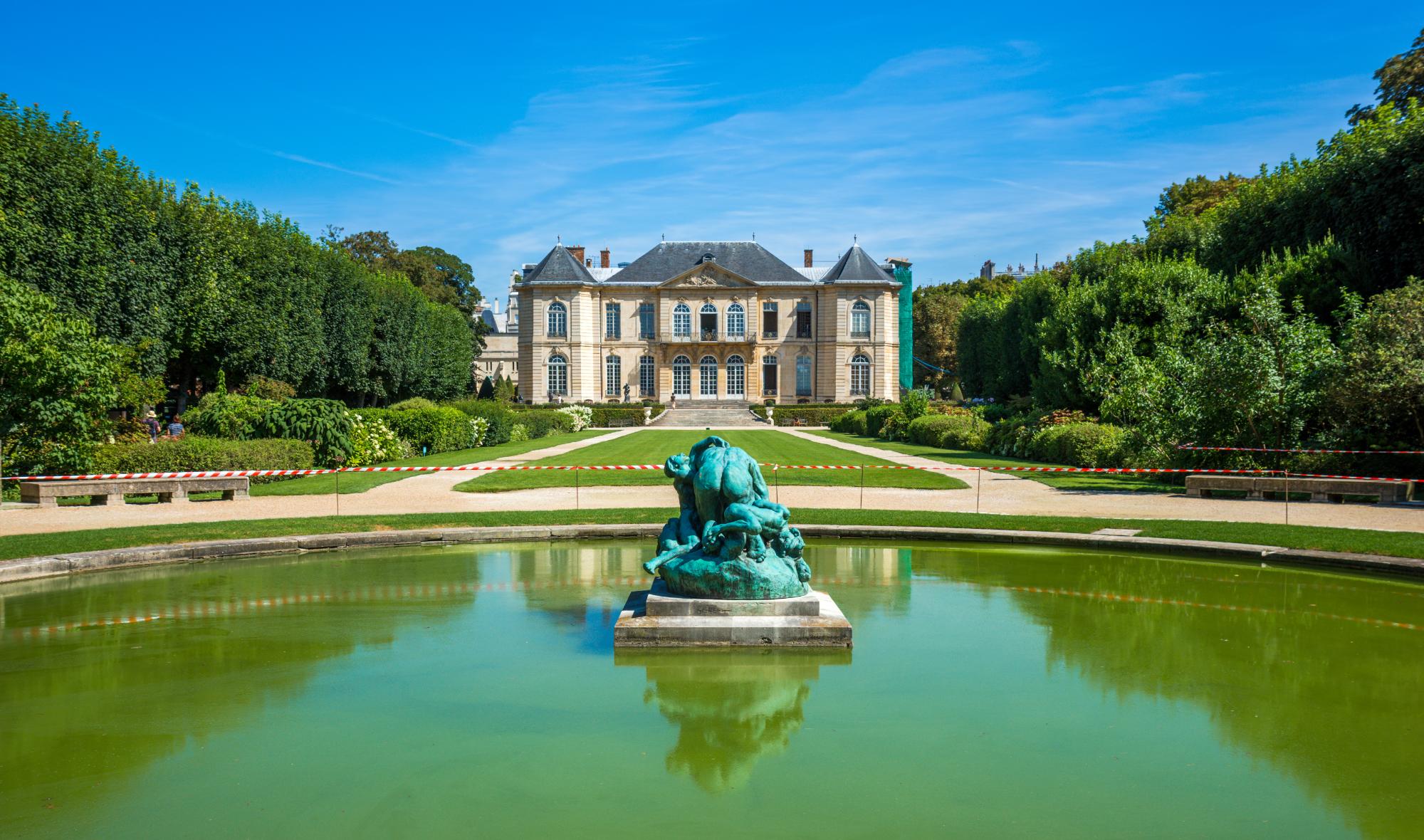 A scenic view of a mansion surrounded by lush gardens with neatly trimmed trees and a reflecting pond featuring a bronze statue.