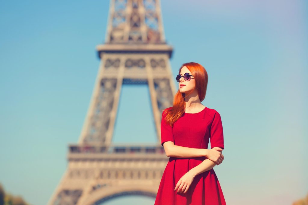 A young woman with long red hair wearing a red dress and sunglasses, standing in front of the Eiffel Tower in Paris.