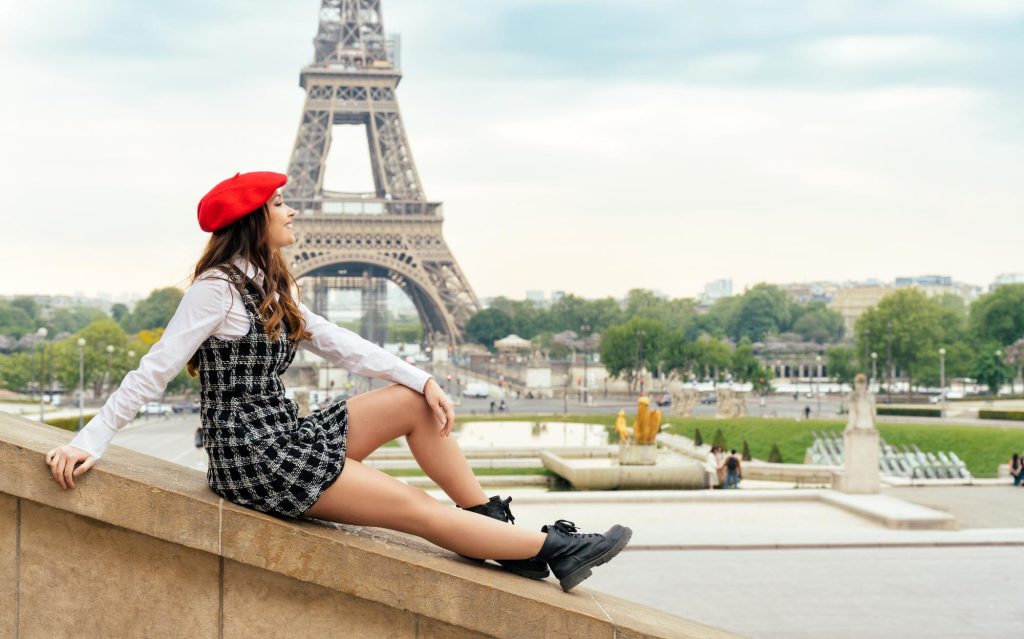 A beautiful young woman sitting gracefully near the Eiffel Tower in Paris, wearing a red beret, a stylish outfit, and enjoying the scenic view.