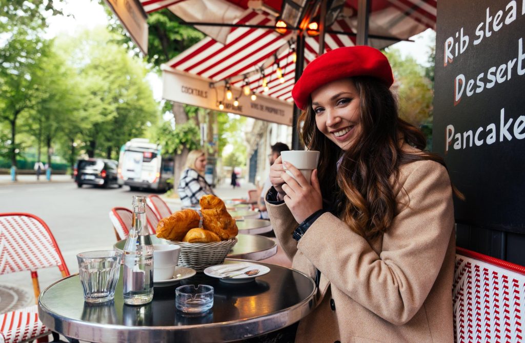 A beautiful young woman in a red beret enjoying coffee and croissants at a cafe in Paris, with trees and street scene in the background.