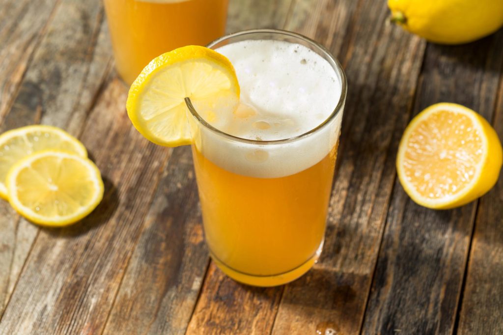 A refreshing glass of beer lemonade garnished with a slice of lemon, served on a wooden table with lemon slices in the background.