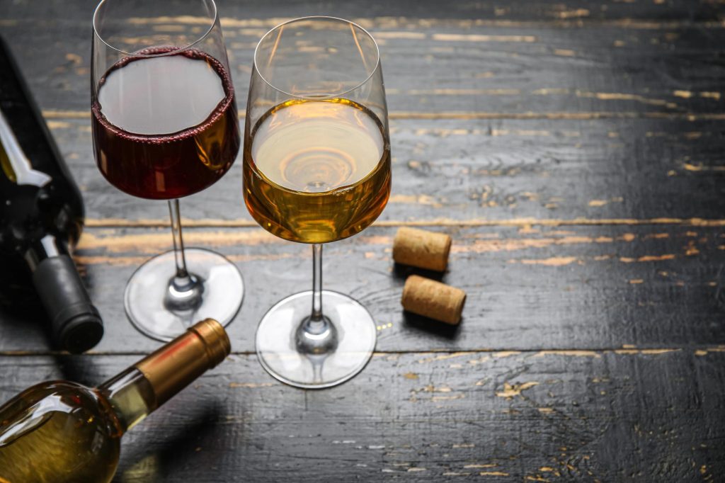 Two wine glasses on a wooden table, one filled with red wine and the other with white wine, accompanied by a bottle of wine and corks.