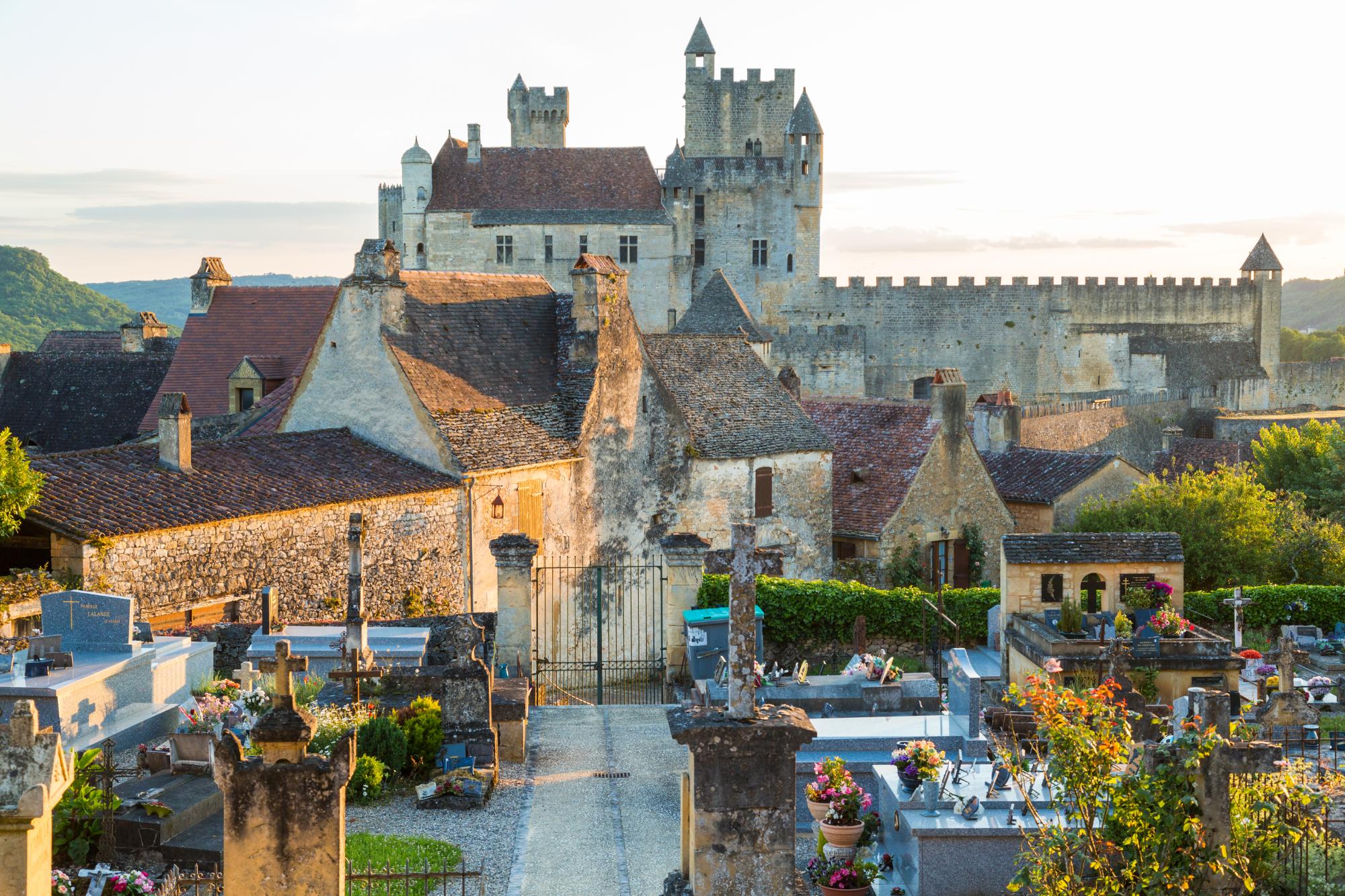 A view of the graveyard in Beynac-et-Cazenac with the historical Beynac Castle in the background during sunset.