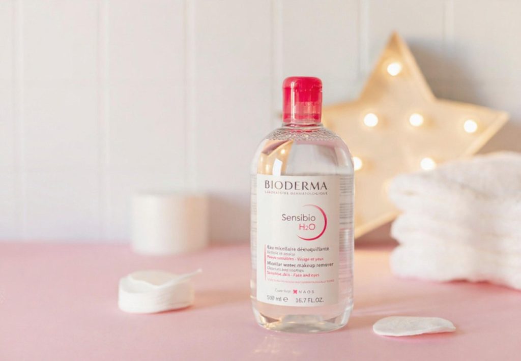 Bioderma Sensibio H2O micellar water bottle on a pink background with cotton pads and a soft towel.