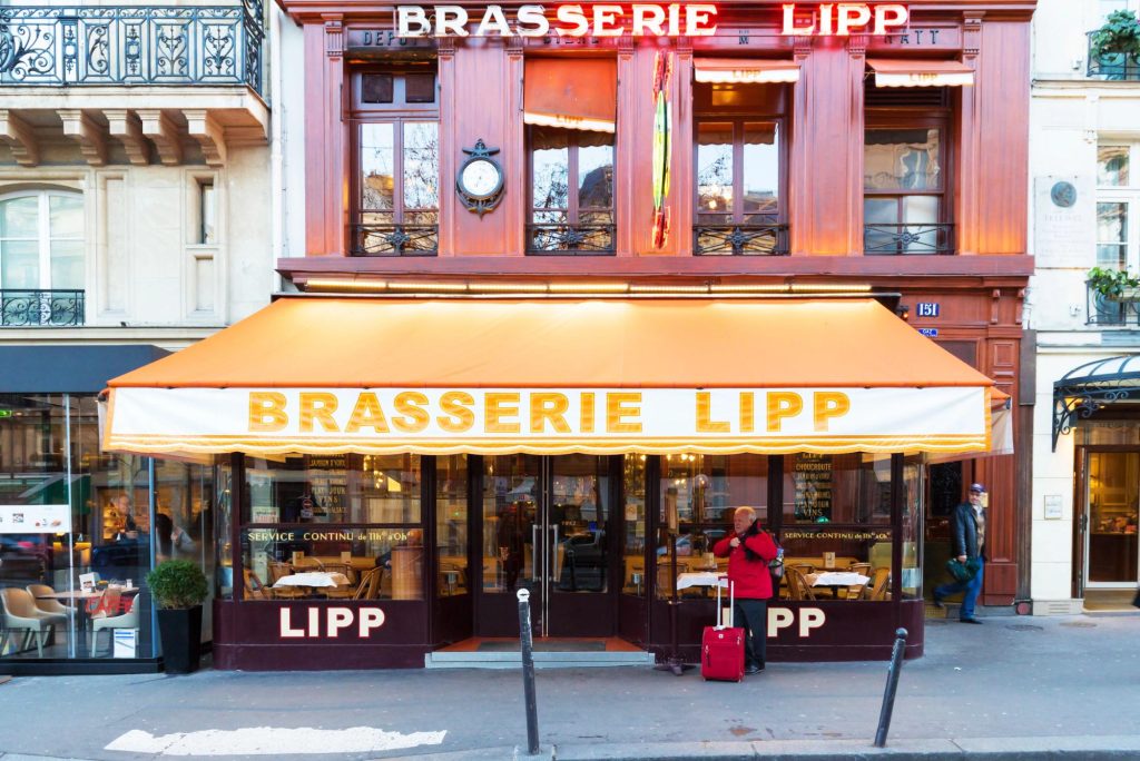 A classic Parisian restaurant facade featuring the name Brasserie Lipp with an orange awning, and a person with a suitcase standing outside.