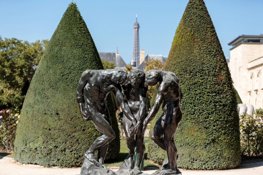 A bronze sculpture depicting three men in a garden, with the Eiffel Tower visible in the background.
