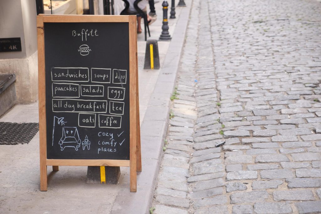 Outdoor chalkboard menu displaying a variety of food and drink options including sandwiches, pancakes, and coffee, with cozy seating symbols.