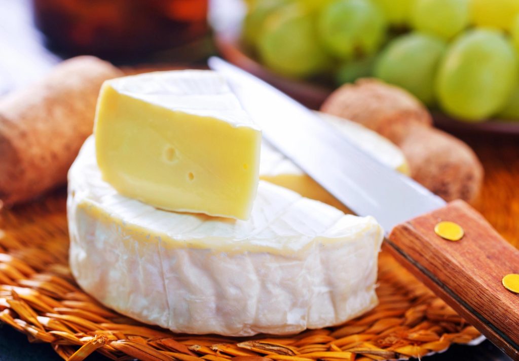 A close-up of a wedge of camembert cheese on a wooden platter, accompanied by a knife and grapes in the background.