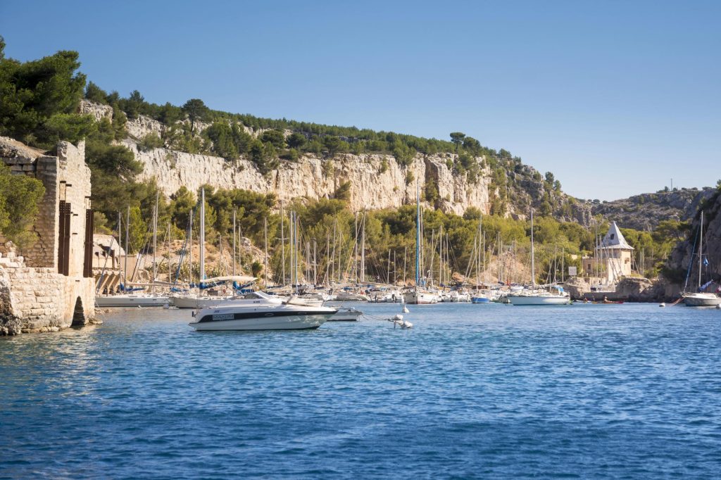 A scenic view of Cassis harbor featuring various sailing boats anchored near rocky cliffs and lush greenery under a clear blue sky.