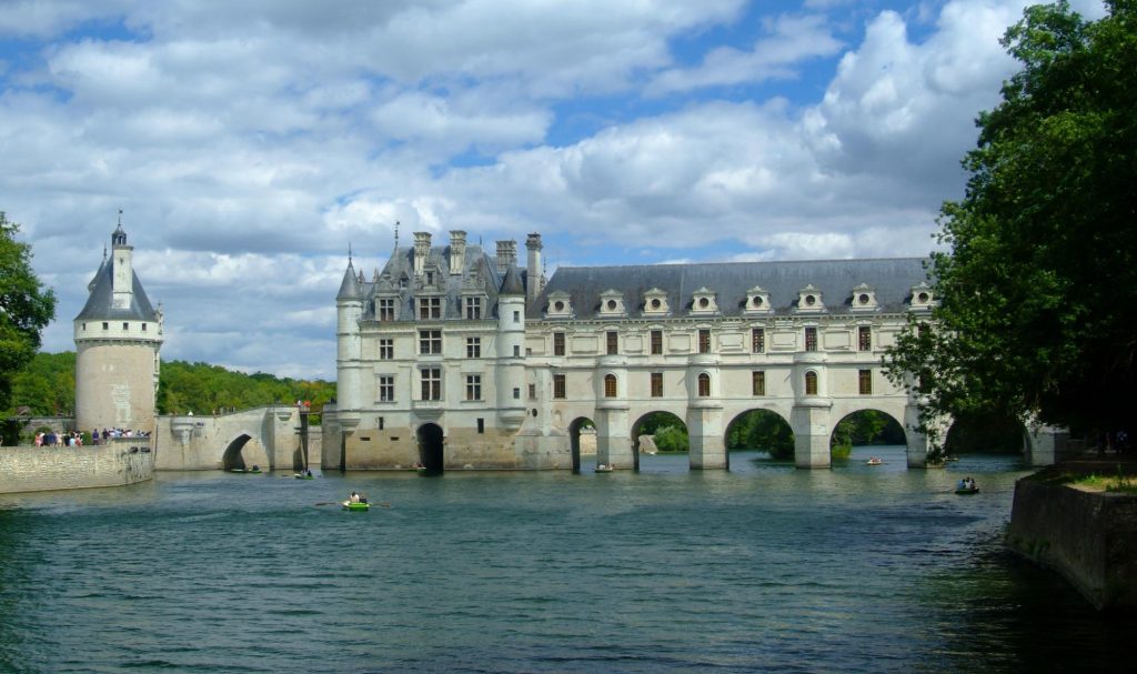 View of Chateau de Chenonceau with its iconic bridge and surrounding water, under a blue sky with clouds.