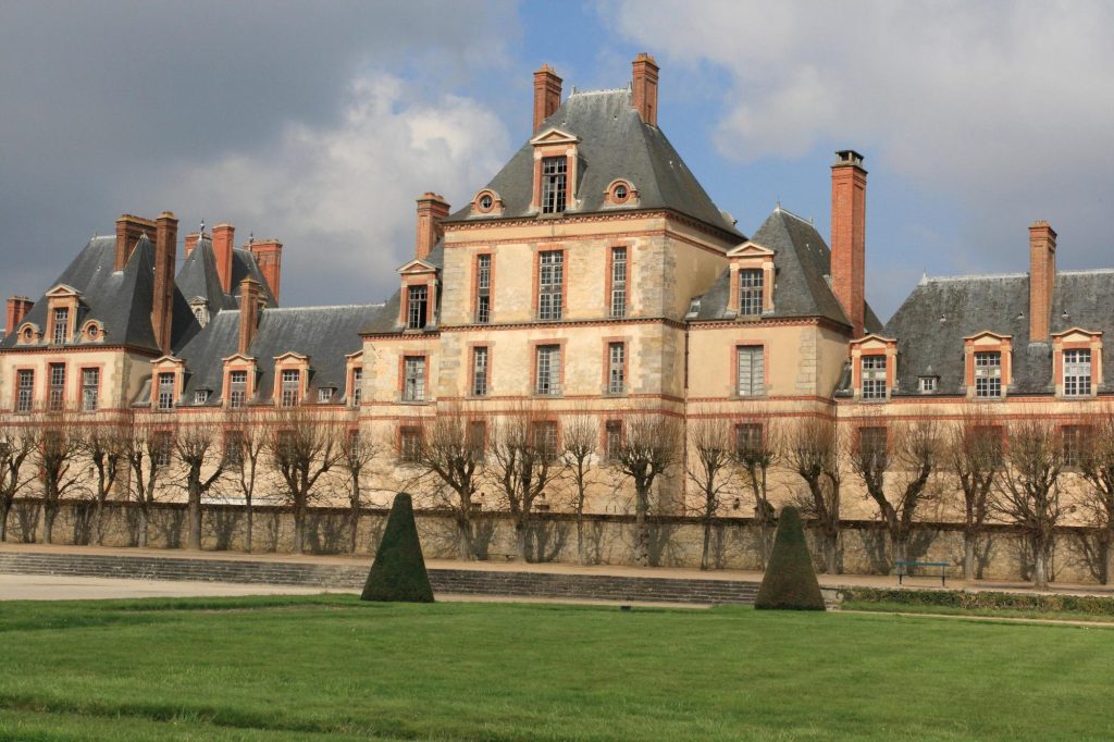 Facade of the Château de Fontainebleau with manicured gardens and topiary in the foreground under a cloudy sky.
