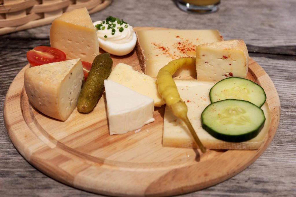 A variety of cheeses including soft, hard, and spreadable types, garnished with cherry tomatoes, pickles, and cucumber slices, served on a wooden platter.