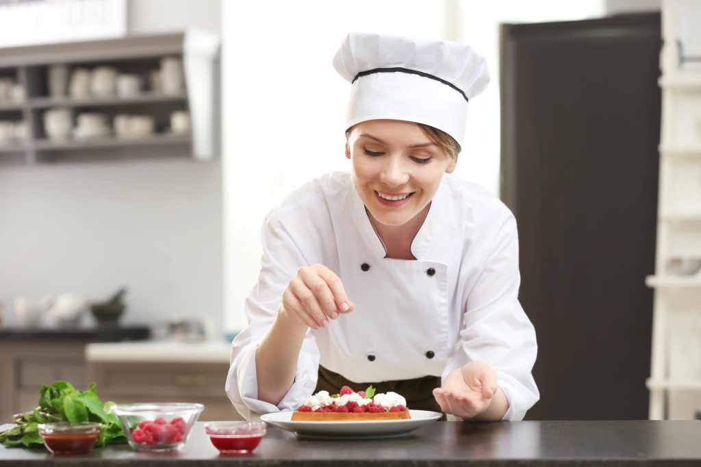 A chef smiling while garnishing a dessert with raspberries and cream in a modern kitchen