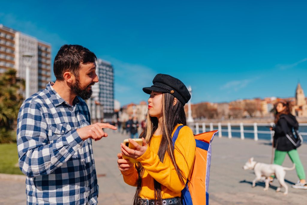 A Chinese woman dressed in an orange sweater and black hat converses with a man in a plaid shirt while holding a smartphone on a sunny day in an urban setting.