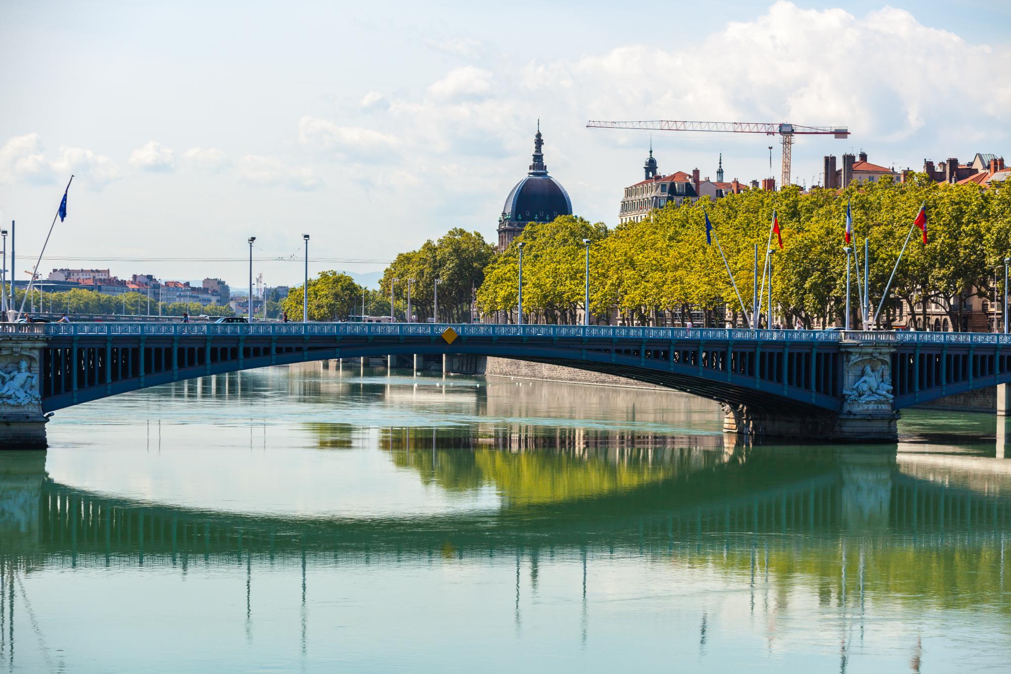 Cityscape of Lyon, France, featuring a bridge with reflections in the water and greenery along the riverbank.