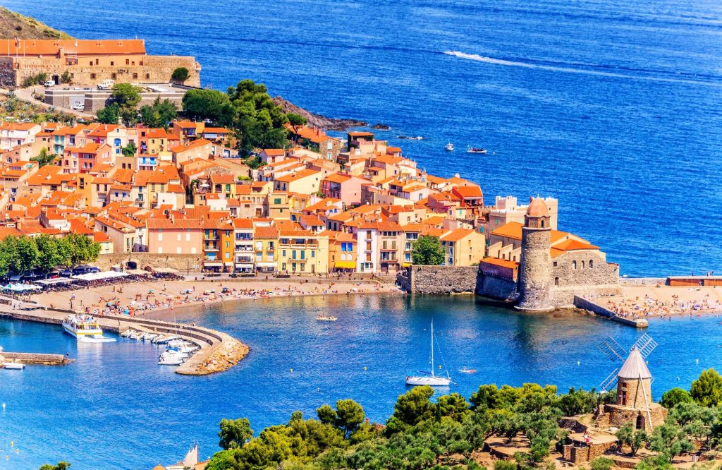 A picturesque view of Collioure, a coastal village in southern France featuring colorful buildings, a sandy beach, and a medieval fortress alongside the blue sea.