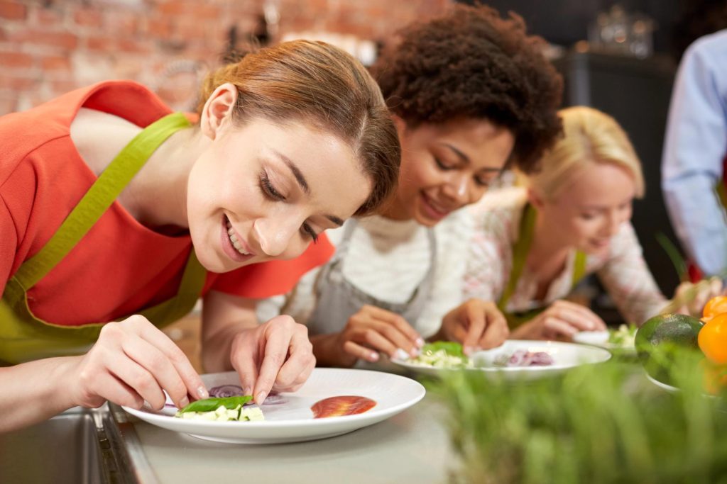 Three women in a cooking class carefully decorating plates with fresh ingredients, showcasing a vibrant and creative culinary experience.