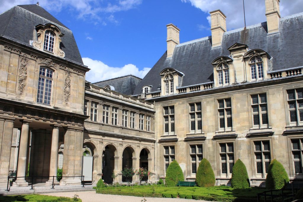 The picturesque courtyard of the Carnavalet Museum in Paris, featuring classical architecture and manicured greenery under a bright blue sky.