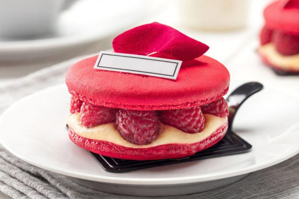 A close-up of a delicious ispahan macaroon filled with raspberries and cream, placed elegantly on a plate, showcasing its vibrant red color and rose petal decoration.