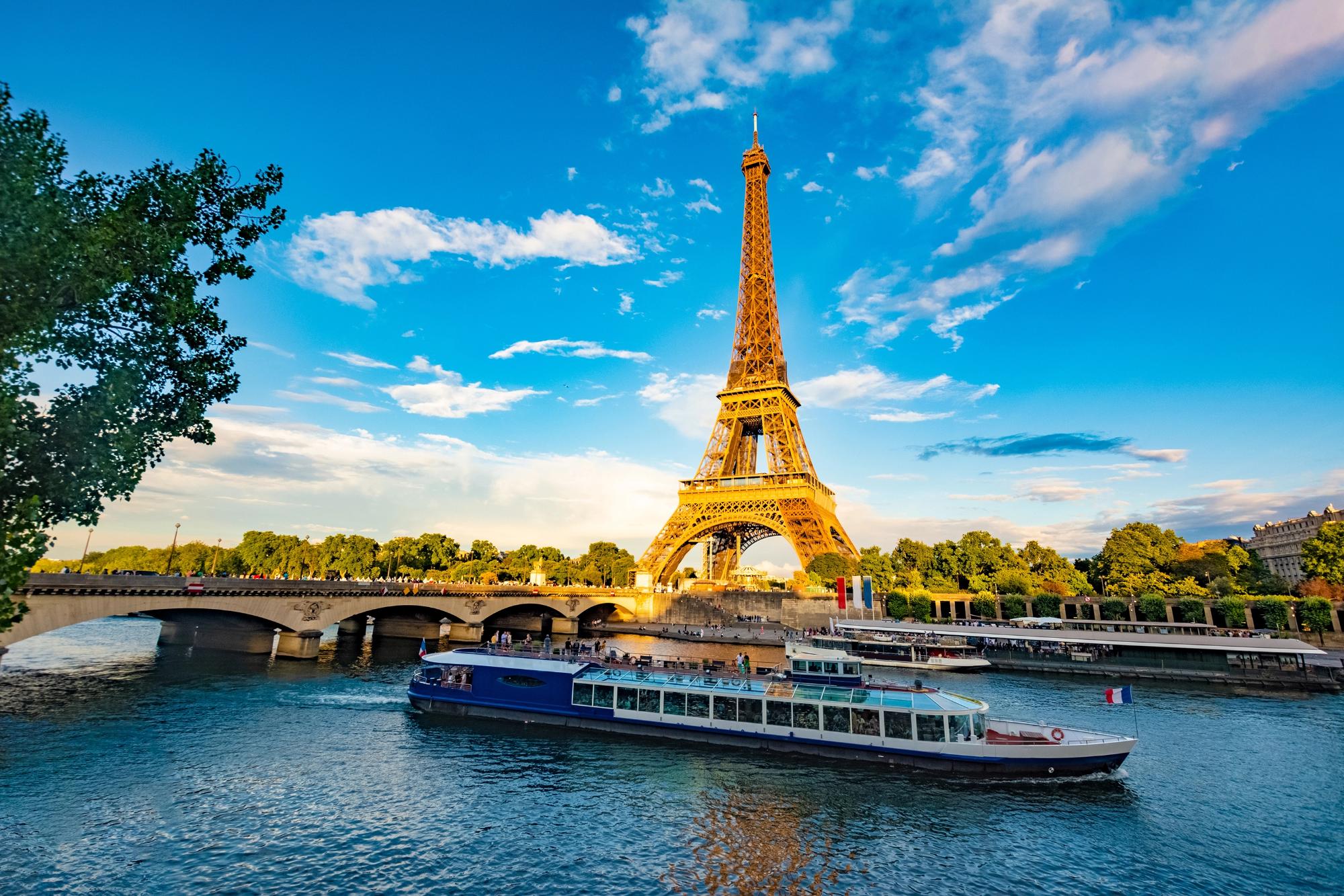 A panoramic view of the Eiffel Tower, the Seine River, and a bridge with boats passing underneath against a blue sky.