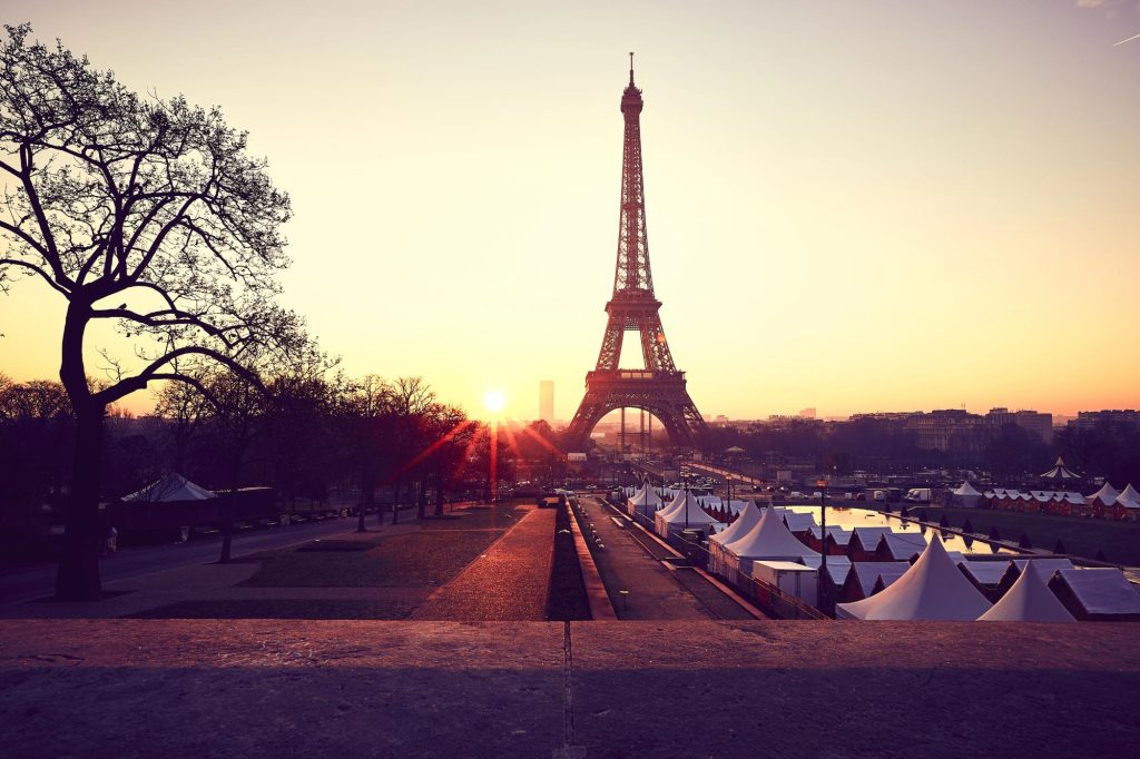 Sunrise view of the Eiffel Tower from Trocadero, with vibrant colors and silhouettes of trees and tents in the foreground.