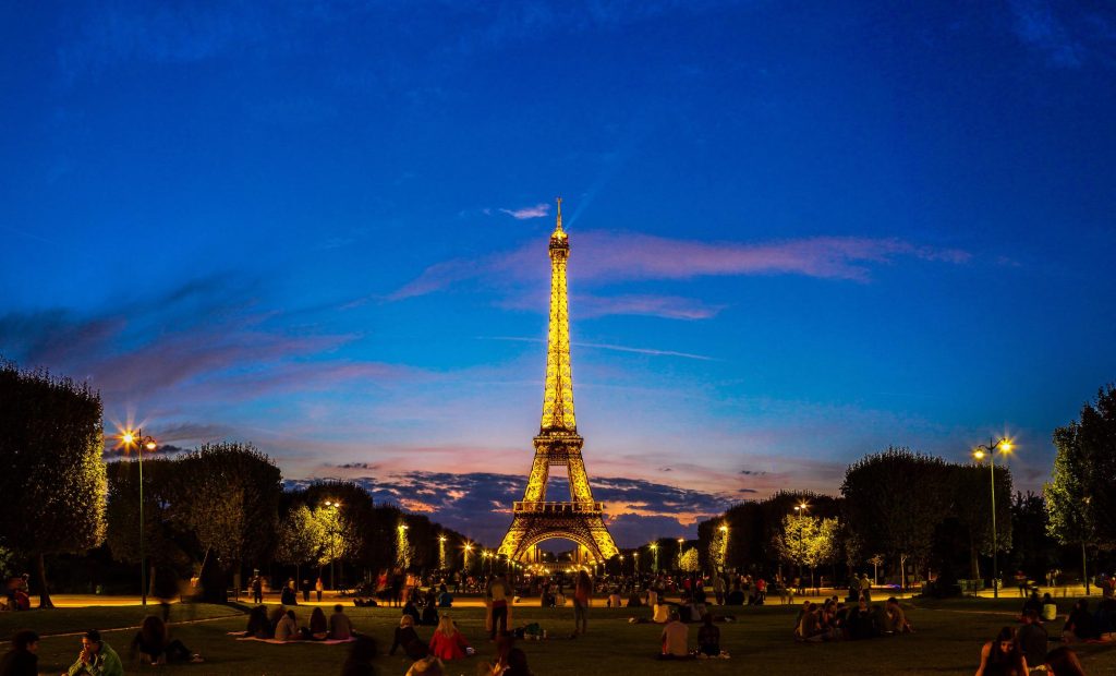 The Eiffel Tower illuminated at sunset in Paris, with people relaxing in the park under a blue sky.