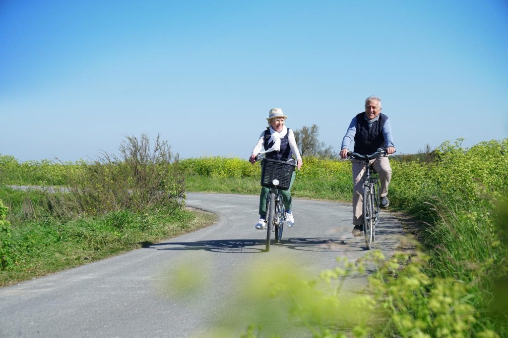 An elderly couple happily cycling on a scenic path surrounded by greenery and wildflowers on a bright sunny day.