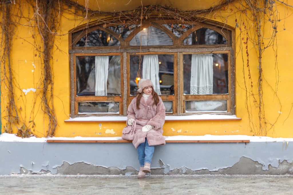 A stylish woman wearing a pink fur coat sits on a bench in front of a yellow wall with wooden windows, surrounded by vines and snow.