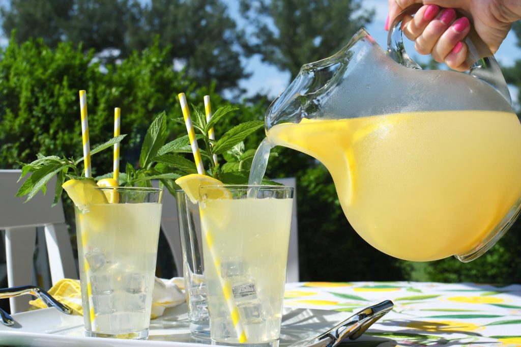 A woman pouring lemonade from a pitcher into tall glasses with lemon slices and mint leaves, enjoying a sunny outdoor setting.