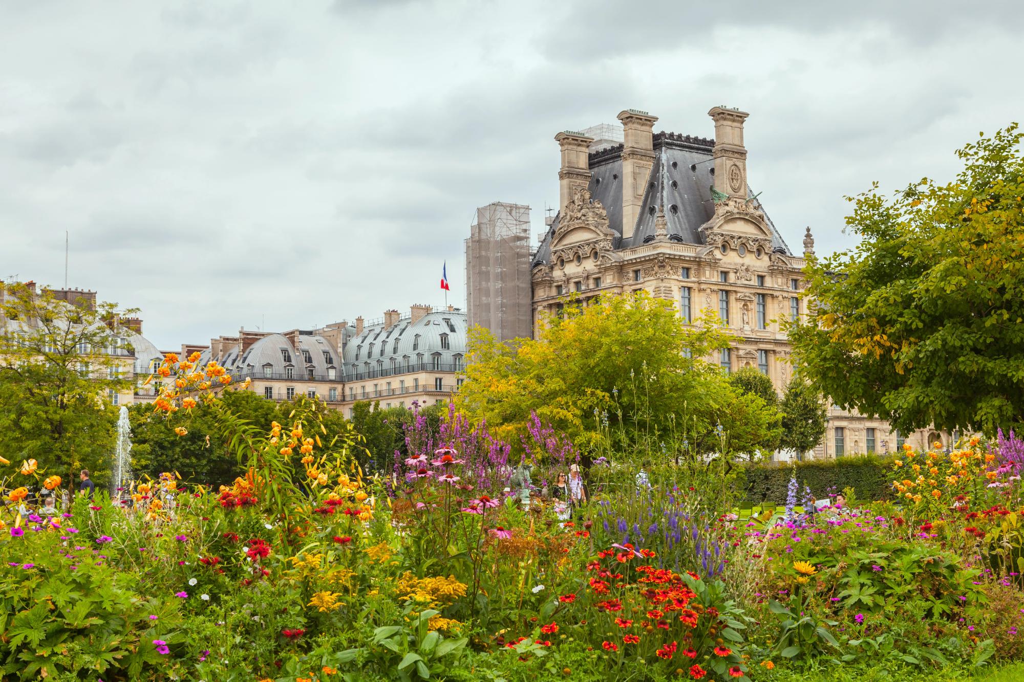 A beautiful garden filled with colorful flowers in the foreground, featuring a historic building with intricate architecture in the background, set against a cloudy sky in Paris.