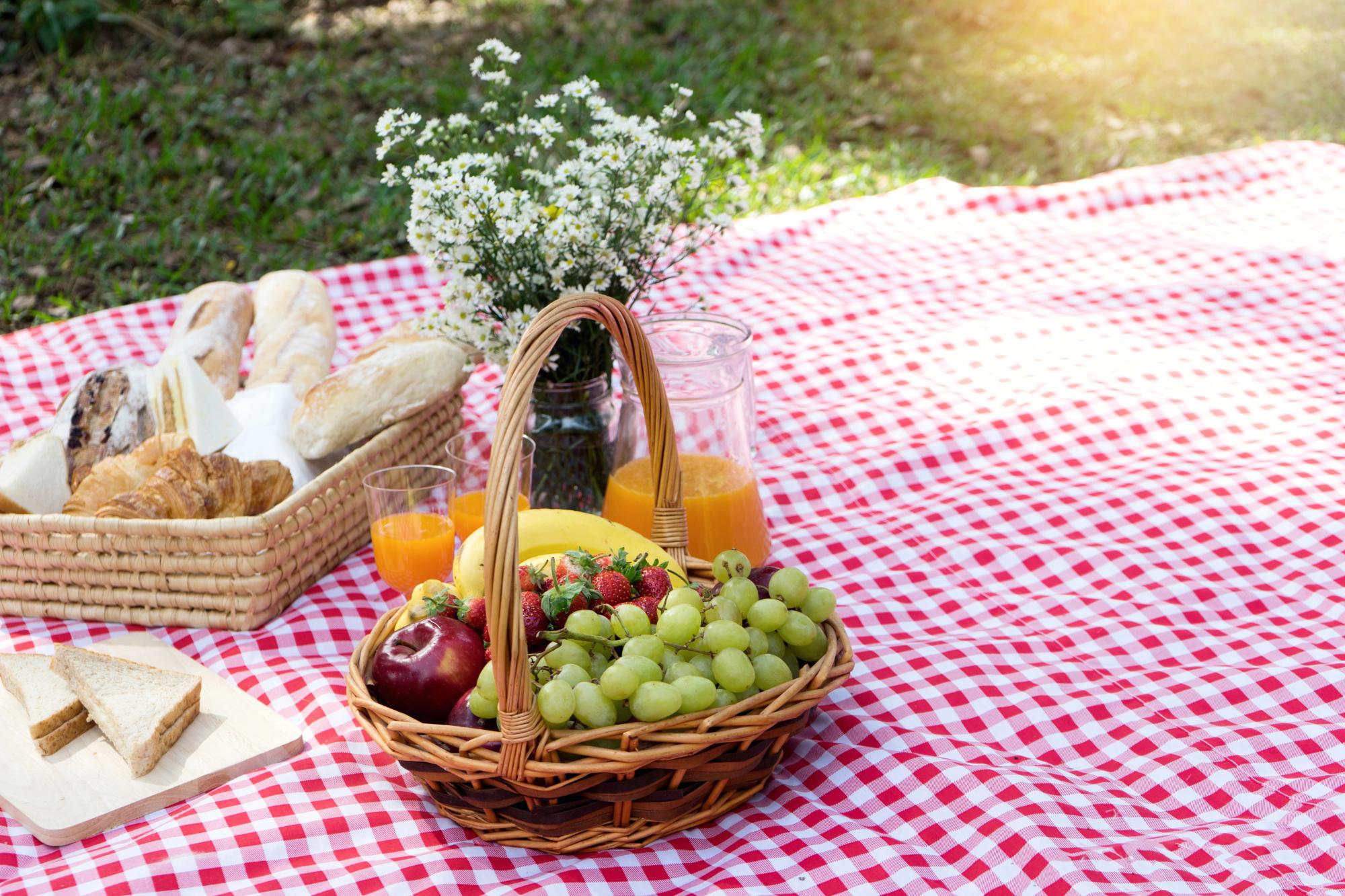 A picnic setup featuring a basket filled with fruits, a variety of breads, and glasses of orange juice on a red and white checkered blanket.