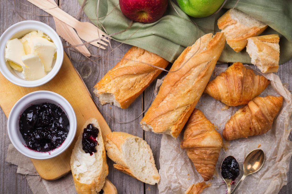 A fresh baguette served with butter and jam on a wooden board, accompanied by croissants and apples on a rustic table.