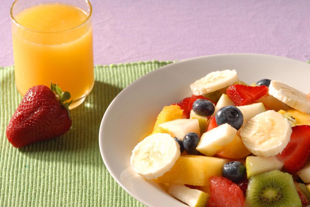 A bowl of fresh seasonal fruit including strawberries, bananas, blueberries, and kiwi, accompanied by a glass of fresh orange juice.