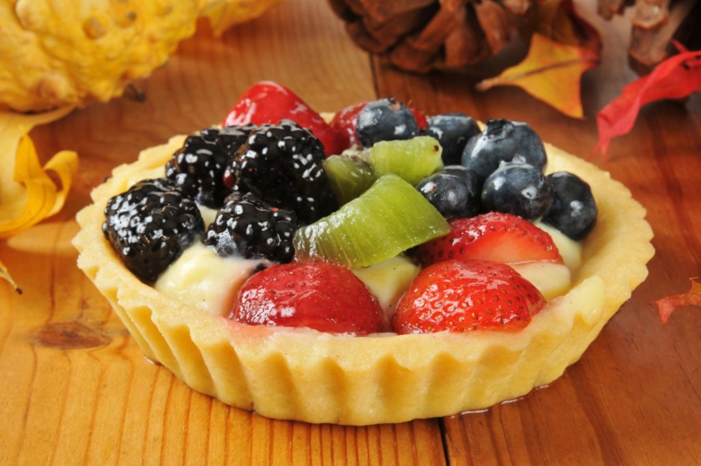 A colorful fruit tart topped with fresh strawberries, blueberries, blackberries, kiwi, and a creamy filling, placed on a wooden surface.