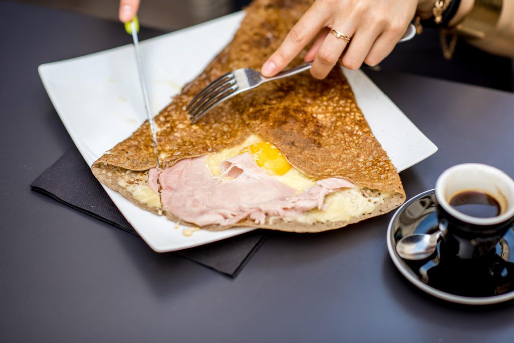 A close-up of a galette filled with ham and a fried egg, being cut with a fork and knife, accompanied by a cup of black coffee.