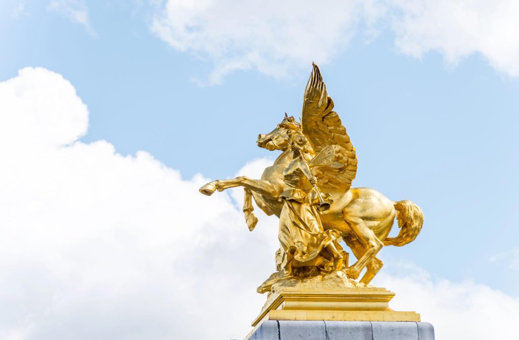 Golden statues on the Pont Alexandre III bridge in Paris, featuring a winged horse and a figure holding a scale and a trumpet against a blue sky.