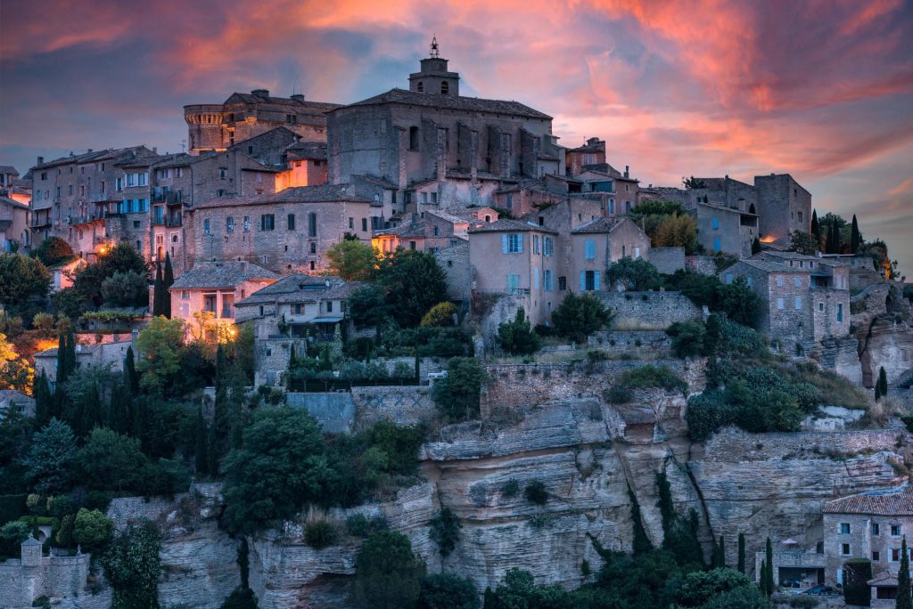 A scenic view of Gordes town in Provence, France, illuminated at twilight, showcasing beautiful stone buildings and a dramatic sky.