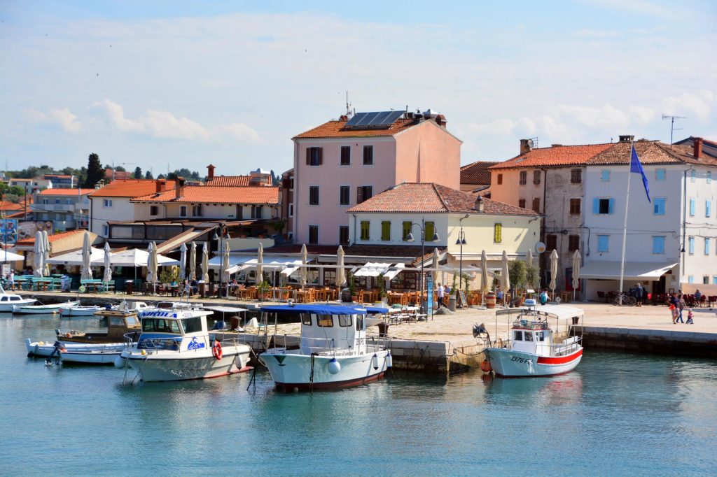A picturesque harbor with several boats docked and colorful buildings lining the waterfront, featuring outdoor dining areas and a clear blue sky.