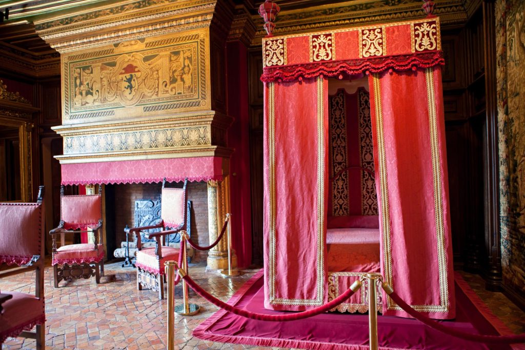 An elegant historical bedroom featuring a red canopy bed, ornate chairs, and a decorated fireplace with intricate designs in a luxurious setting.