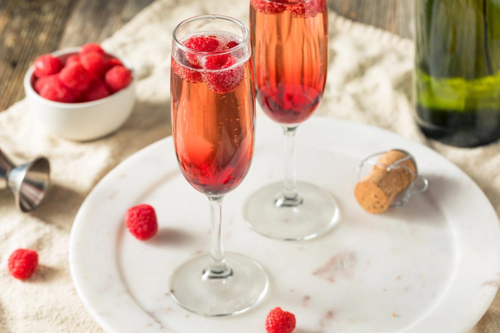 Two sparkling glasses of Kir and Kir Royale cocktails garnished with raspberries, placed on a marble plate with a bottle and cork in the background.
