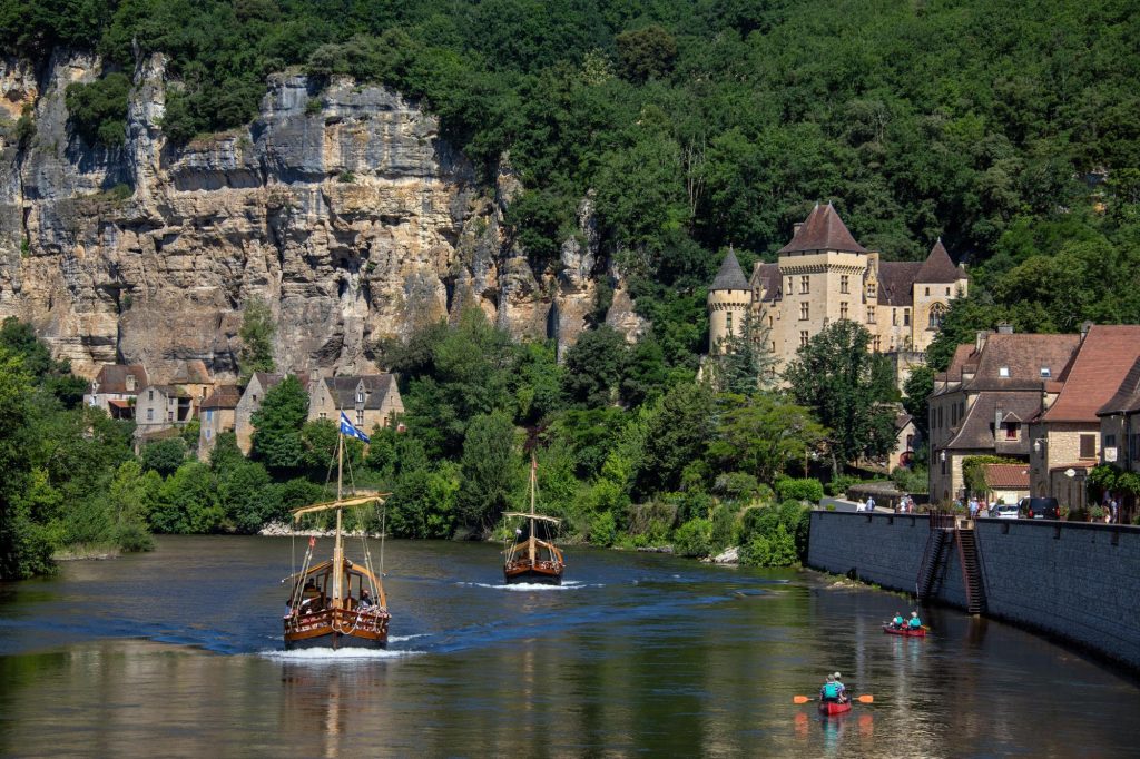 Scenic view of La Roque-Gageac in Dordogne, France, featuring traditional boats on the river, historic buildings along the shore, and lush greenery in the background.